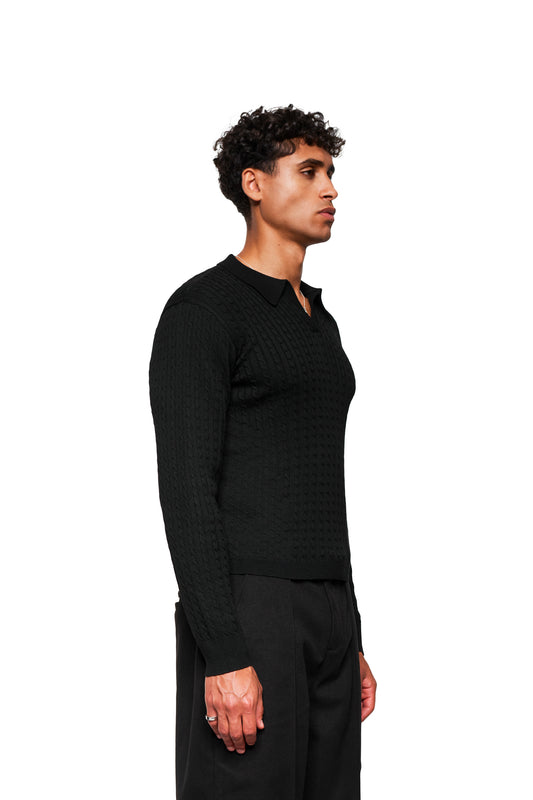 Model trägt Exquisite Cable Knit Polo Black mit Zopf-Strickmuster, eng anliegender Passform, doppeltem Kragen links, gestricktem Bund und Ärmelbündchen.