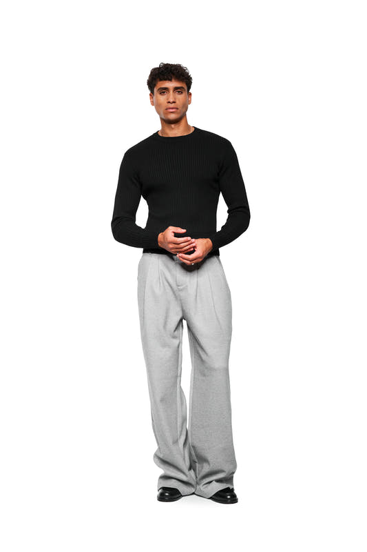 Young man in Exquisite Knit Longsleeve Black, tight-fitting with different knit patterns, combined with wide-cut, flowing gray pants and black leather shoes.