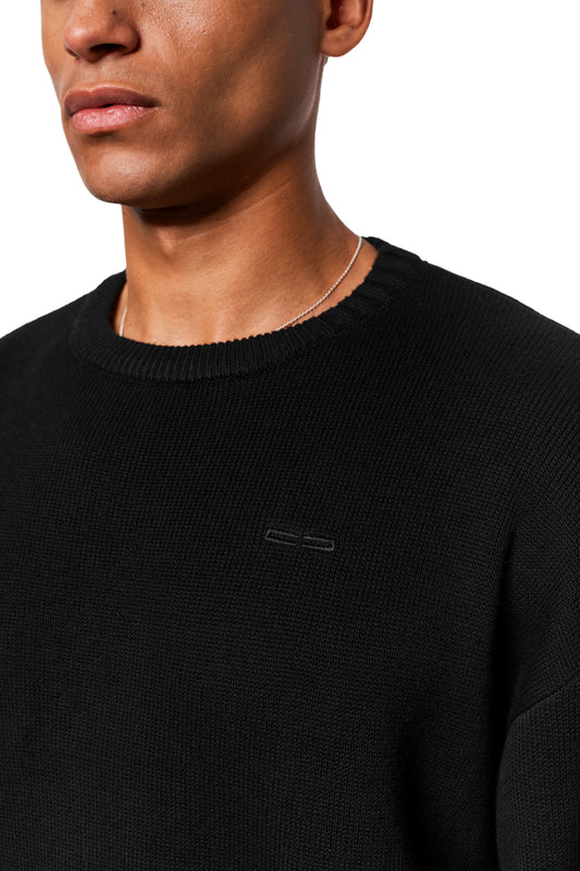 Man wears Exquisite Knit Sweater Black with embroidered OLAKALA icon on the chest; loose fit, various knit patterns on the collar and cuffs.