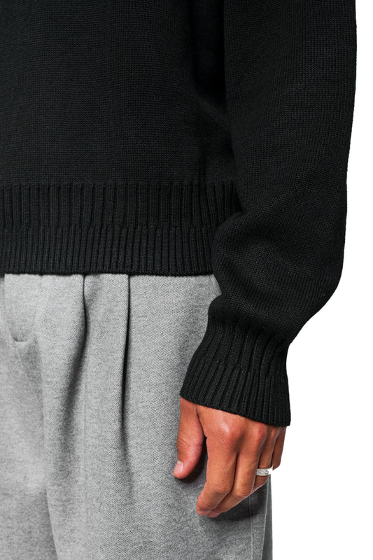 Close-up of a unisex Exquisite Knit Sweater Black with embroidered OLAKALA Icon, knitted cuffs and loose fit, worn with gray pants and silver ring.