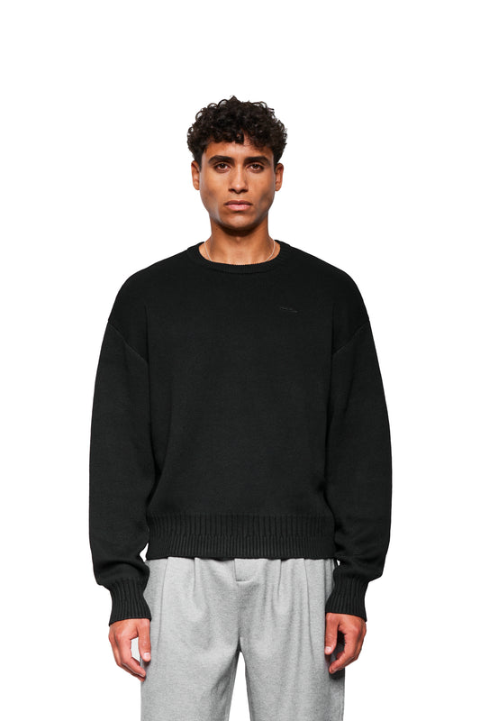Männliches Model trägt Exquisite Knit Sweater Black mit gesticktem OLAKALA Icon, lockerer Passform und verschiedenen Strickmustern an Kragen, Bund und Ärmelbündchen.