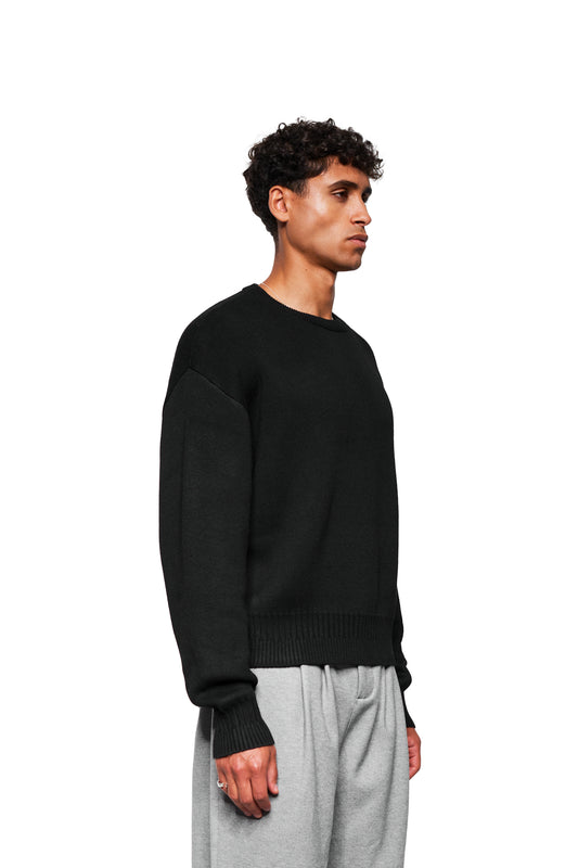 Mann in Profil trägt schwarzen Exquisite Knit Sweater mit gesticktem OLAKALA-Icon und verschiedenen Strickmustern, kombiniert mit grauer Jogginghose. Unisex und locker geschnitten.