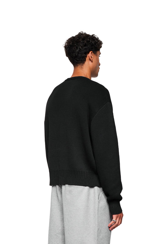 Young man wearing an Exquisite Knit Sweater Black with OLAKALA icon and unique knit patterns, loose fit, seen from behind, slight profile visible.