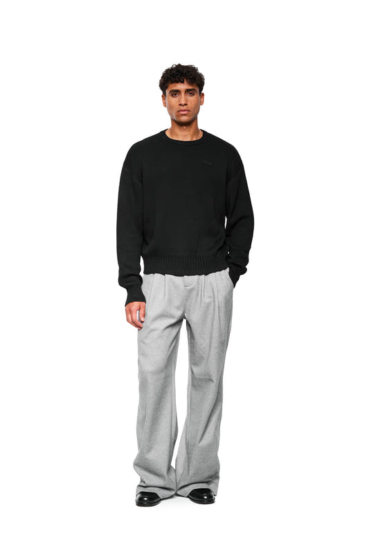 Young man wears Exquisite Knit Sweater Black with OLAKALA Icon and ribbed waistband, combined with loose-fitting trousers and leather shoes, emphasizing casual style.