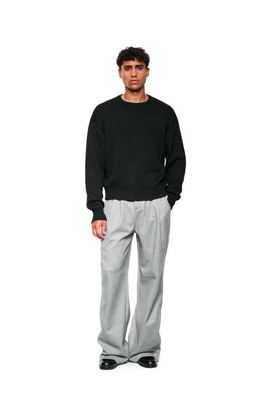 Young man wears Exquisite Knit Sweater Black with OLAKALA Icon and ribbed waistband, combined with loose-fitting trousers and leather shoes, emphasizing casual style.