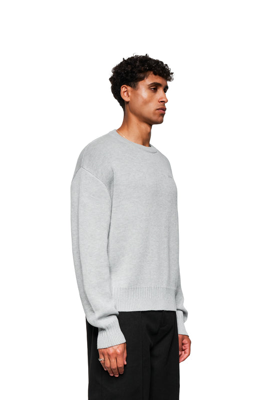 Junger Mann in grauem Exquisite Knit Sweater mit gesticktem OLAKALA-Icon, lockerer Passform, angestricktem Bund und variierenden Strickmustern, seitlich stehend.