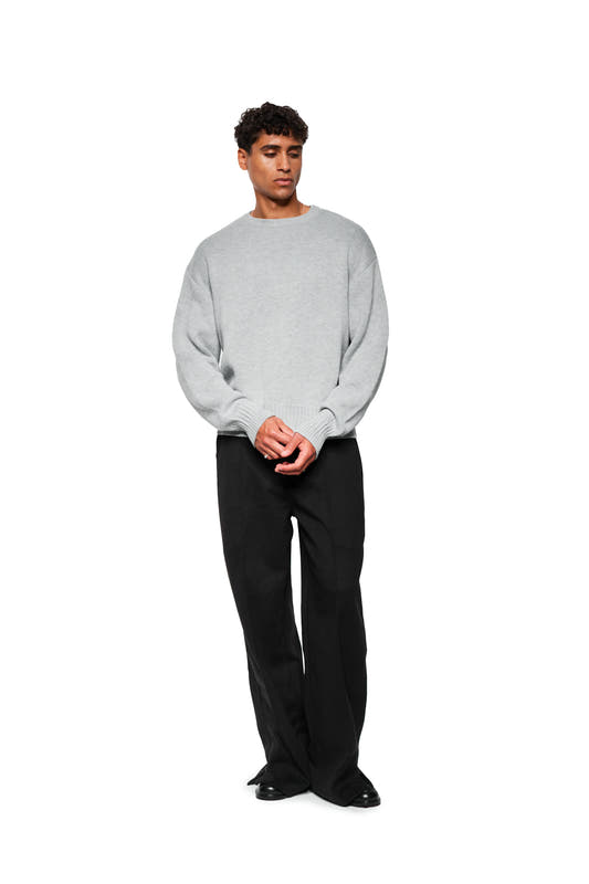 Male model wears Exquisite Knit Sweater Gray with embroidered OLAKALA icon, ribbed cuffs and loose cut, combined with black pants and shoes.