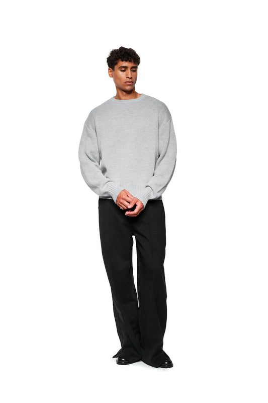 Male model wears Exquisite Knit Sweater Gray with embroidered OLAKALA icon, ribbed cuffs and loose cut, combined with black pants and shoes.