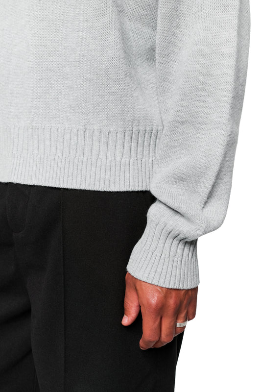 Person wears Exquisite Knit Sweater Gray with embroidered OLAKALA Icon, different knit patterns on cuffs and collar, loose fit, unisex, silver ring visible on finger.
