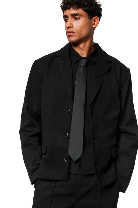 Männliches Modell trägt ein schwarzes Ensemble mit Monogram Tie Black, einer schwarzen Krawatte mit Monogramm, und blickt zur Seite.