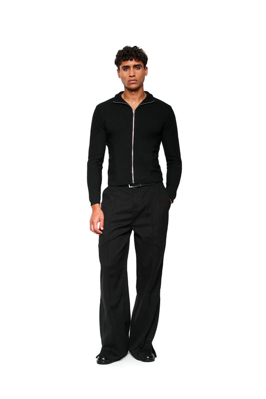 Man wears Exquisite Knit Jacket Black with an anatomical knit pattern, stand-up collar and close fit, combined with wide-cut pants and elegant shoes.
