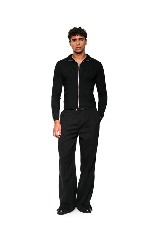 Man wears Exquisite Knit Jacket Black with an anatomical knit pattern, stand-up collar and close fit, combined with wide-cut pants and elegant shoes.