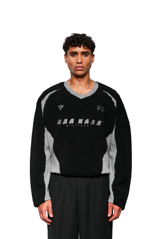 Junger Mann trägt Fleece V-Neck Sweater Black mit gesticktem OLAKALA Logo und Icons, V-Ausschnitt, elastischem Saum, unisex oversized Passform.