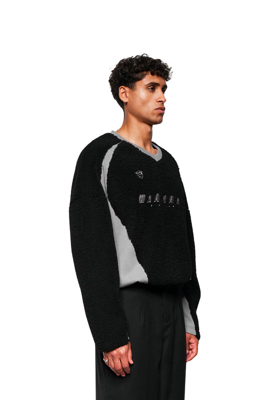 Männliches Model im Profil mit Fleece V-Neck Sweater Black, zeigt gesticktes OLAKALA Logo und Icons, kombiniert mit schwarzer Hose und silberner Halskette.
