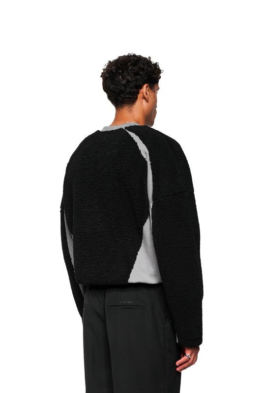 Rückansicht eines Mannes im Fleece V-Neck Sweater Black mit Oversize-Schnitt und gesticktem OLAKALA-Logo, über einem grauen Shirt und schwarzen Hosen.