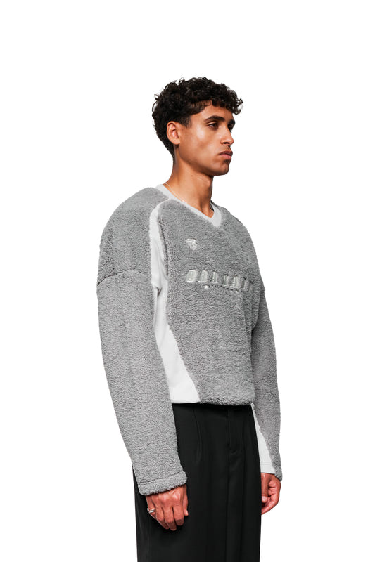 Junger Mann im Profil trägt einen grauen Fleece V-Neck Sweater von OLAKALA, mit gesticktem Logo und Icons, kombiniert mit schwarzen Hosen.