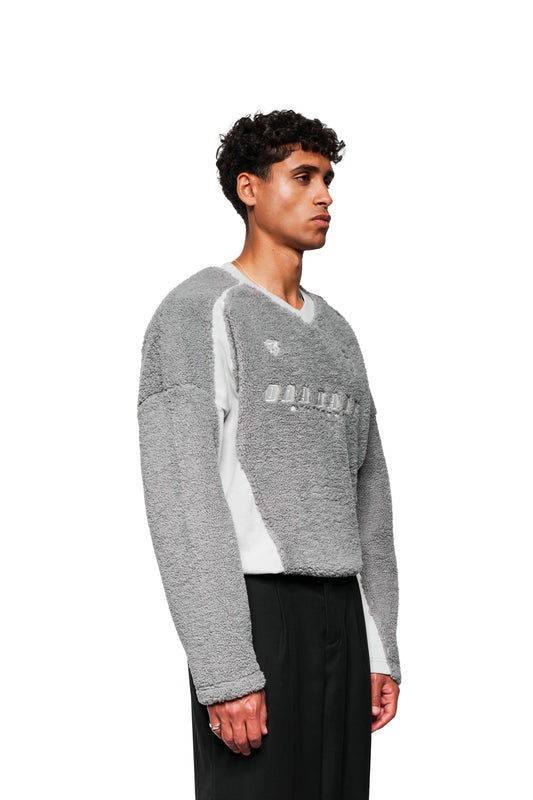 Junger Mann im Profil trägt einen grauen Fleece V-Neck Sweater von OLAKALA, mit gesticktem Logo und Icons, kombiniert mit schwarzen Hosen.