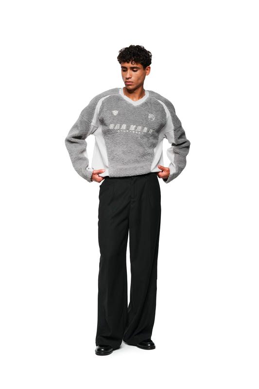 Junger Mann trägt Fleece V-Neck Sweater Grau mit gesticktem OLAKALA Stuttgart Logo, sportlichen Icons und verstellbarem Saum, kombiniert mit schwarzen Hosen und Lederschuhen.