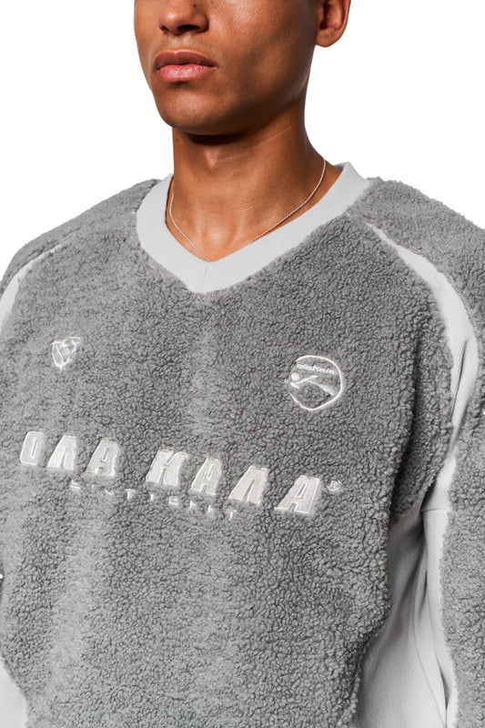 Mann trägt einen grauen Fleece V-Neck Sweater mit ONA KAA-Schriftzug, gestickten Logos und verstellbarem Saum, passend zur OLAKALA Unisex-Kollektion.