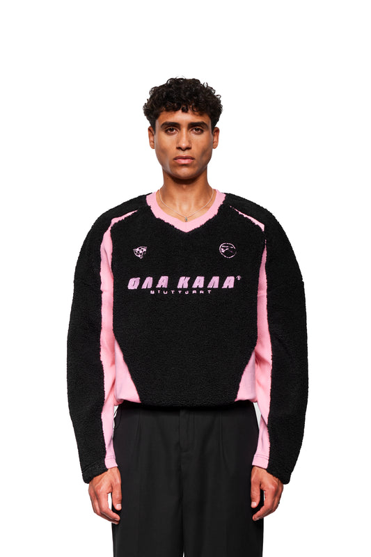 Männliches Model trägt den Fleece V-Neck Sweater Pink mit gesticktem OLA KALA Stuttgart-Logo und Icons auf der Brust, kombiniert mit schwarzen Hosen.