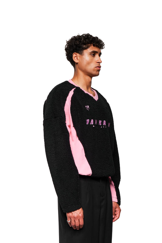 Männliches Model im Profil trägt schwarzen Fleece V-Neck Sweater Pink mit rosa Einsätzen, gesticktem OLAKALA Stuttgart Logo, und Icons. Kombiniert mit rosa Hemd und schwarzen Hosen.