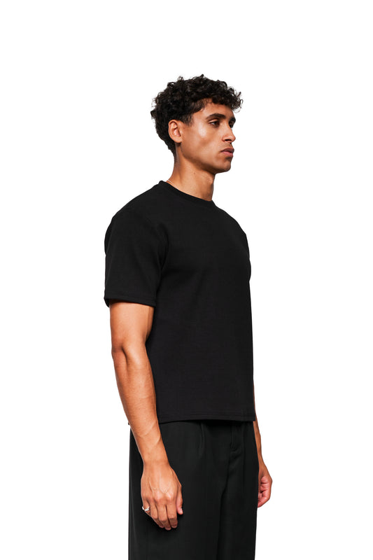 Junger Mann im Profil trägt das Exquisite Rib T-Shirt Black in lockerer Passform aus festem Rippstoff, präsentiert in einem studio-beleuchteten Setting. Unisex-Design.