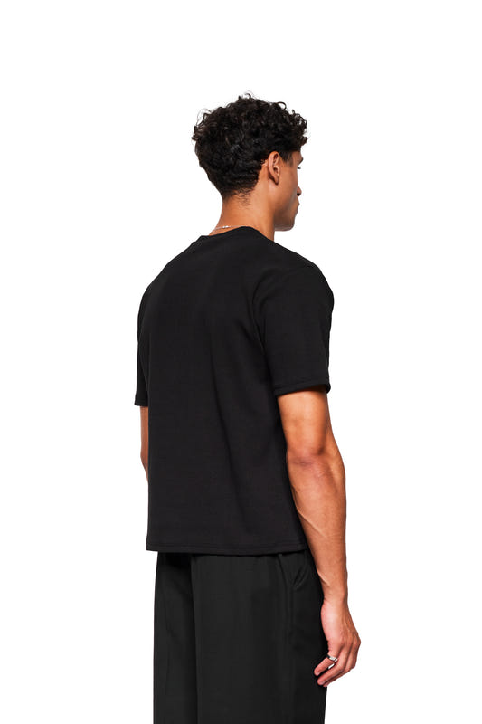 Rückansicht eines Mannes im Exquisite Rib T-Shirt Black, locker geschnitten, unisex, mit kurzen Ärmeln, aus festem Rippstoff, passend zu weiten schwarzen Hosen.