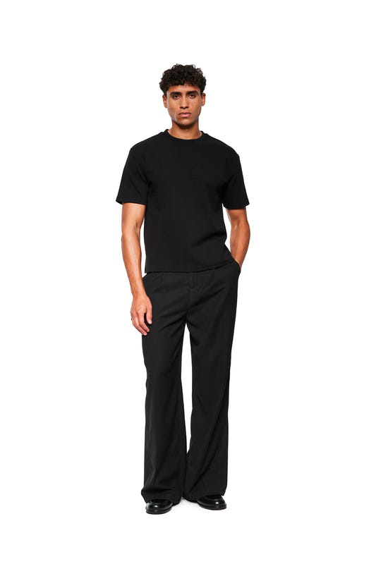 Junger Mann in Exquisite Rib T-Shirt Black, lockerem Schnitt, kombiniert mit weiten schwarzen Hosen und eleganten Lederschuhen, präsentiert minimalistischen Stil.