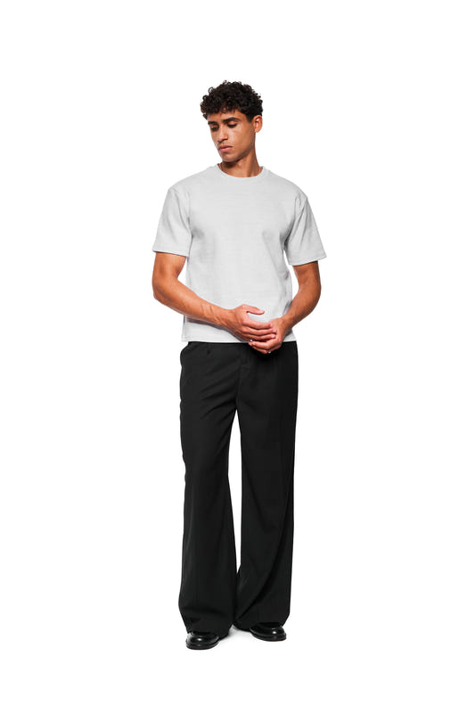 Junger Mann trägt Exquisite Rib T-Shirt Gray Melange, locker über weiten schwarzen Hosen und schwarzen Schuhen, Hände vor dem Körper gefaltet. Unisex, lockere Passform.