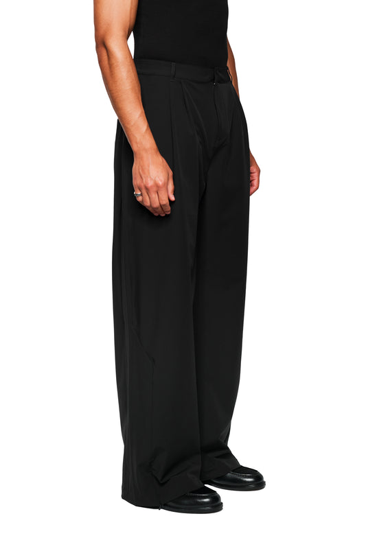 Person trägt Exquisite Tech Pants Black mit weitem Bein und Bundfalten, kombiniert mit schwarzen Loafers. Sichtbare Details: versetzte Naht, OLAKALA Icon über Gesäßtasche.