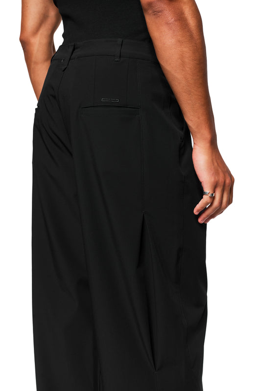 Person wears Exquisite Tech Pants Black with loose fit, pleats, offset side seam and embroidered OLAKALA icon. Hand visible with silver rings.