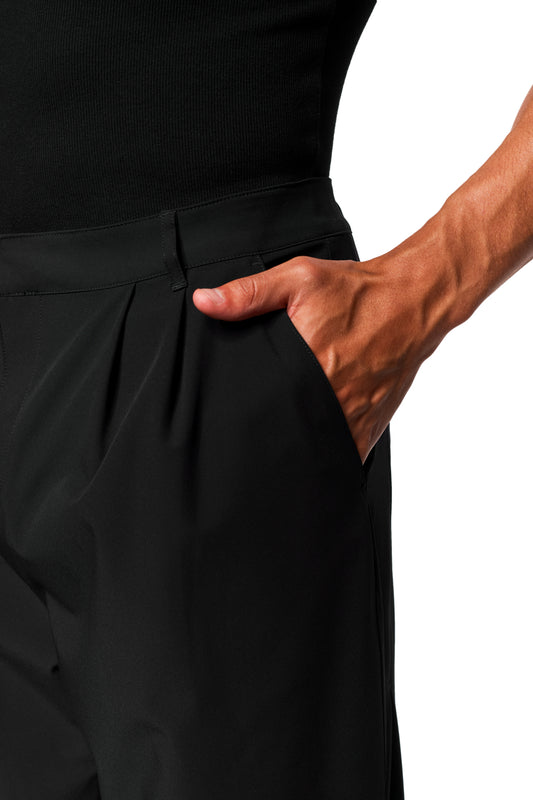 Person wears Exquisite Tech Pants Black with asymmetrical pocket opening, pleats and loose fit. Hand casually in the side pocket, presenting robust, stretchy material.