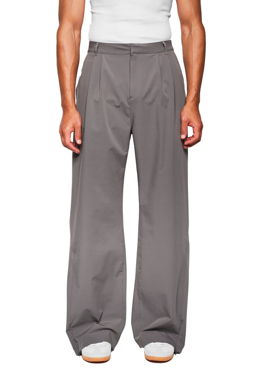 Person trägt Exquisite Tech Pants Gray mit weitem Bein, asymmetrischem Tascheneingriff und Bundfalten; weiße Sneakers und weißes Topabschluss erkennbar.