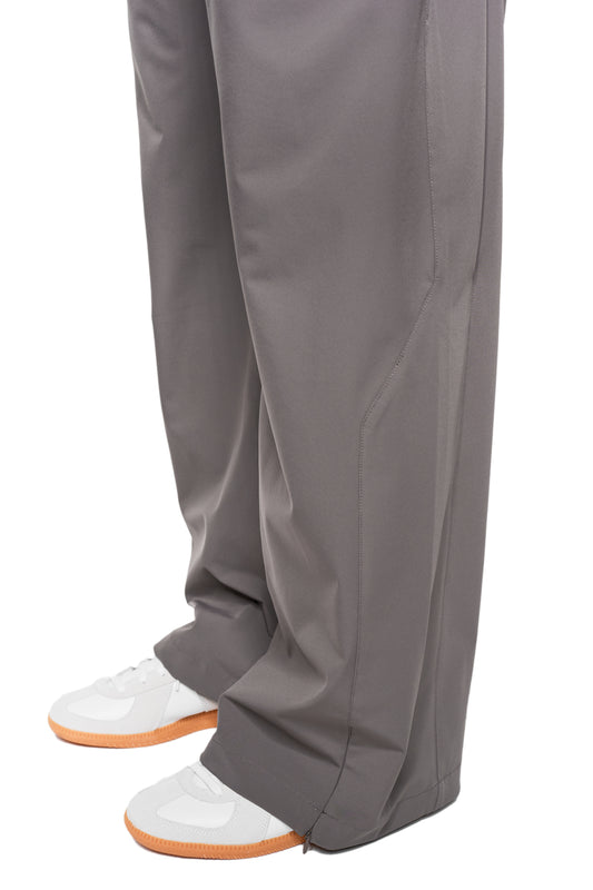 Exquisite Tech Pants Gray: unisex trousers with asymmetrical pocket opening, pleats and embroidered OLAKALA icon, combined with white sneakers on a white background.