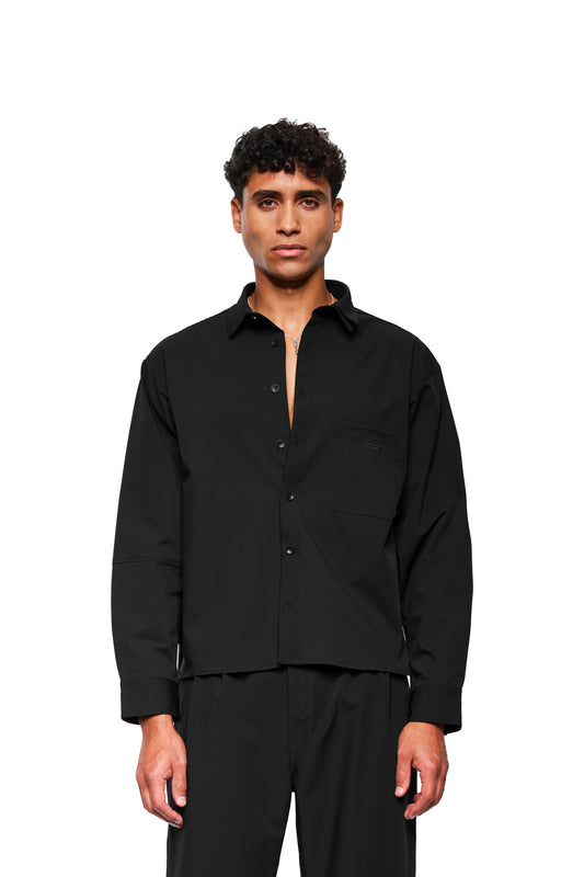 Ein Model trägt das Exquisite Tech Shirt Black mit doppeltem Kragen und Brusttasche mit OLAKALA Stickerei, kombiniert mit schwarzen Hosen, lockere Passform.