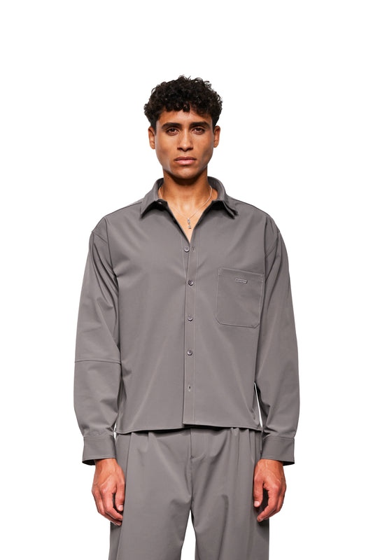 Person trägt das Exquisite Tech Shirt Gray mit doppeltem Kragen, Brusttasche mit OLAKALA Stickerei und abgerundetem Saum, kombiniert mit passenden grauen Hosen.