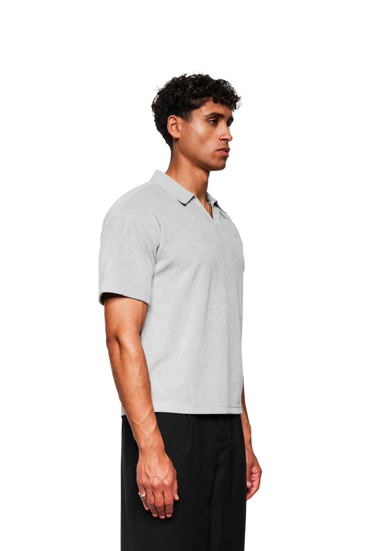 Junger Mann im Profil trägt ein Terry Polo Gray aus strukturiertem Frotteematerial mit Logo-Stickerei auf der Brust und kombiniert mit lockeren schwarzen Hosen.