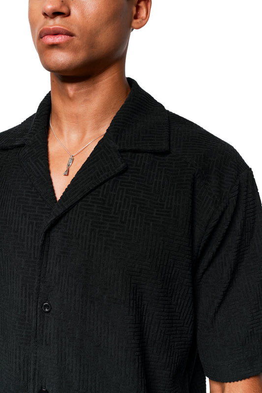 Person in einem kurzärmeligen, schwarzen Terry-Hemd mit 3D-Monogramm-Effekt und Safari-Kragen, trägt eine silberne Kette mit kleinem Anhänger.