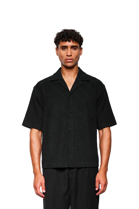 Junger Mann trägt ein schwarzes Terry Kurzarmhemd mit 3D-Monogramm-Effekt und Safari-Kragen, passende Hose, vor weißem Hintergrund.