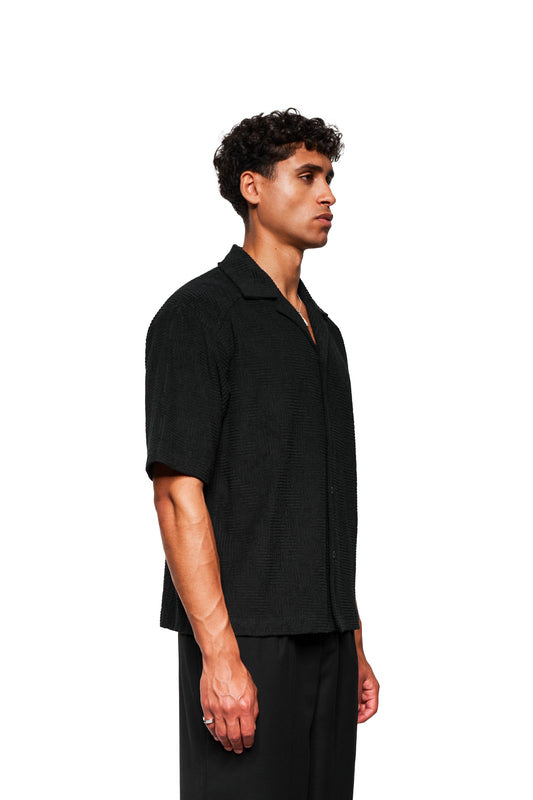 Junger Mann im Profil mit weichem, strukturiertem Frottee-Kurzarmhemd, Safari-Kragen und 3D-Monogramm-Effekt, passend zur OLAKALA Unisex-Kollektion.
