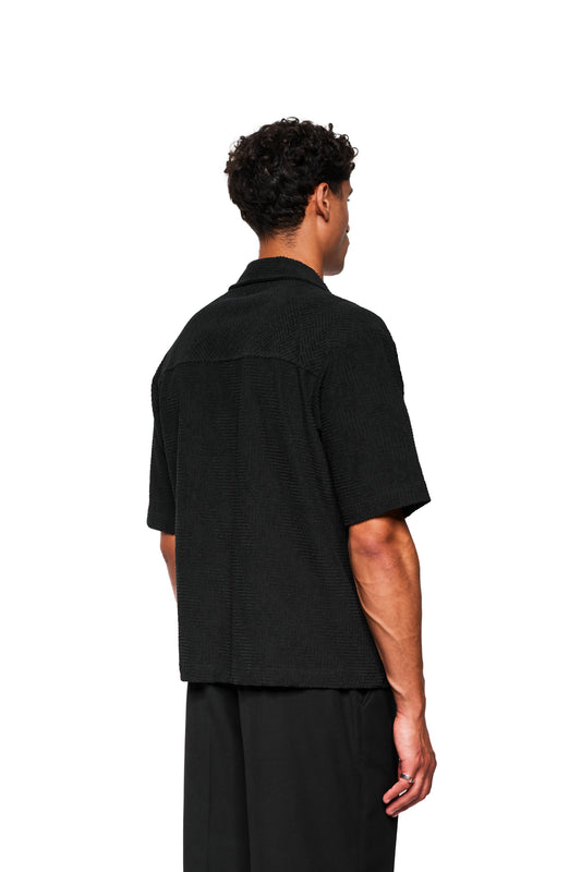 Mann von hinten in lockerem, schwarzem Terry Kurzarmhemd aus strukturiertem Frottee mit Safari-Kragen und 3D-Monogramm, präsentiert von OLAKALA.