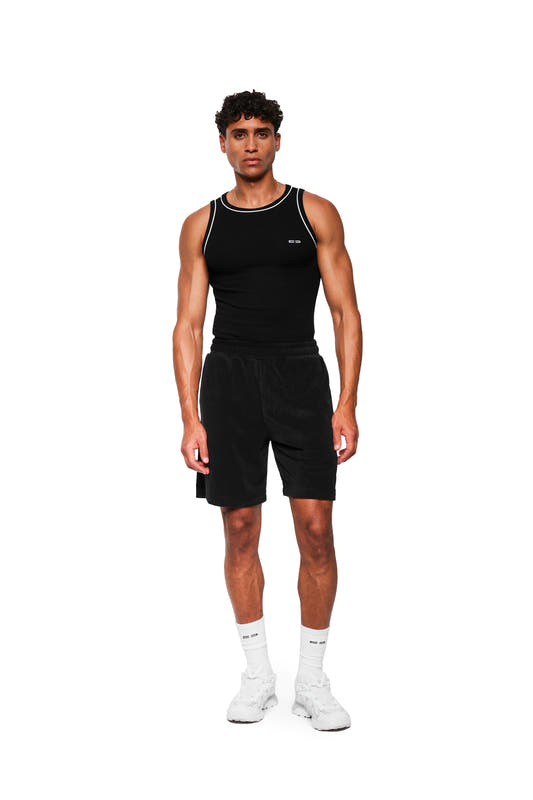 Junger Mann trägt schwarze Terry Shorts mit elastischem Bund, Kordelzug und Logo-Stickerei, kombiniert mit ärmellosem Tanktop und Sneakers.