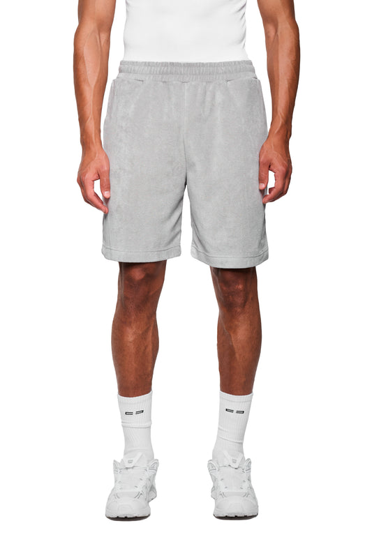Männermodel trägt graue Terry Shorts mit elastischem Bund, Seitentaschen und Logo-Stickerei, kombiniert mit weißen Crew-Socken und Chunky-Sneakers.