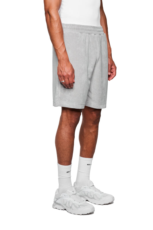 Person trägt Terry Shorts Gray aus weichem Frottee mit elastischem Bund und Logo-Stickerei, kombiniert mit weißen Socken und Turnschuhen.
