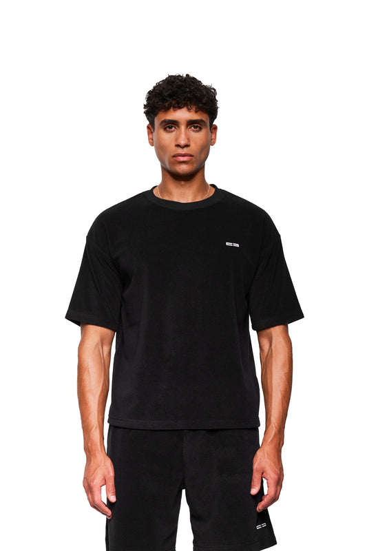 Männliches Model in einem schwarzen Terry T-Shirt mit minimalistischer weißer Logo-Stickerei auf der Brust, kombiniert mit passenden Shorts. Entspanntes, lässiges Design von OLAKALA.