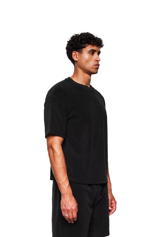 Junger Mann trägt ein Terry T-Shirt Black aus weichem Frotteematerial mit Logo-Stickerei auf der Brust, präsentiert in einem minimalistischen Modestil.