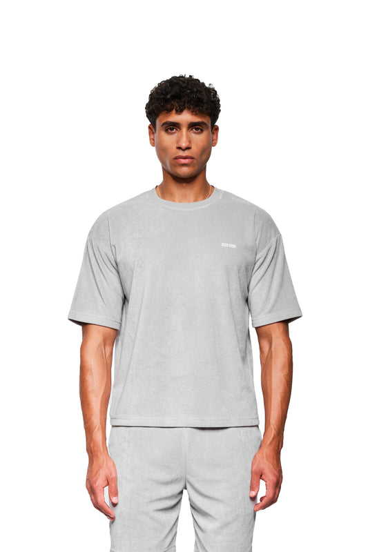 Männliches Model in einem grauen Terry T-Shirt von OLAKALA, aus weichem Frotteematerial, mit Logo-Stickerei auf der Brust, präsentiert lässigen, unisex Look.