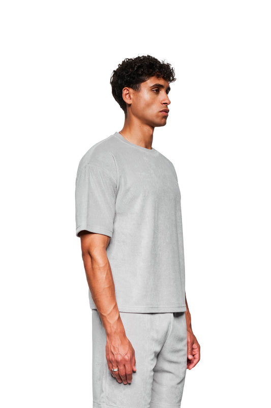Männliches Modell in Profilansicht trägt ein graues Terry T-Shirt mit Logo-Stickerei auf der Brust und passenden Shorts, ergänzt durch eine silberne Kette.