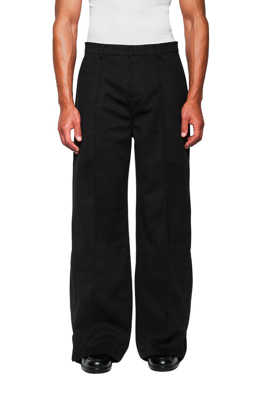 Modell trägt Exquisite Dress Pants Black mit weitem Bein und gesticktem OLAKALA-Icon, Biese vorn, seitlichem Reißverschluss am Saum, lockerer Unisex-Passform.