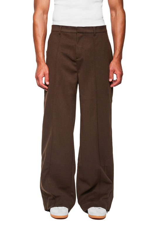 Exquisite Dress Pants Brown getragen von Modell, mit weitem Schnitt, Bundfalten, versetzter Naht, Reißverschluss am Saum und OLAKALA-Icon über der Gesäßtasche.